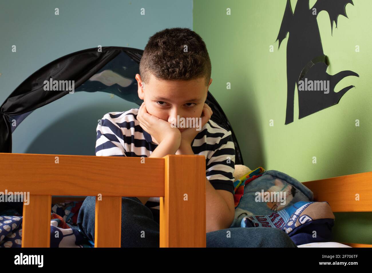 9 year old boy unhappy in bedroom after conflict with parent, resting chin on palms, sitting bed Stock Photo