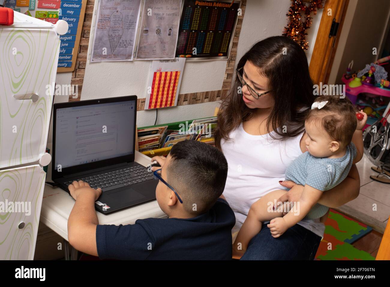7 year old boy, wearing glasses using laptop computer for school work, mother assisting him holding her 7 month old sister, learning  at home Stock Photo