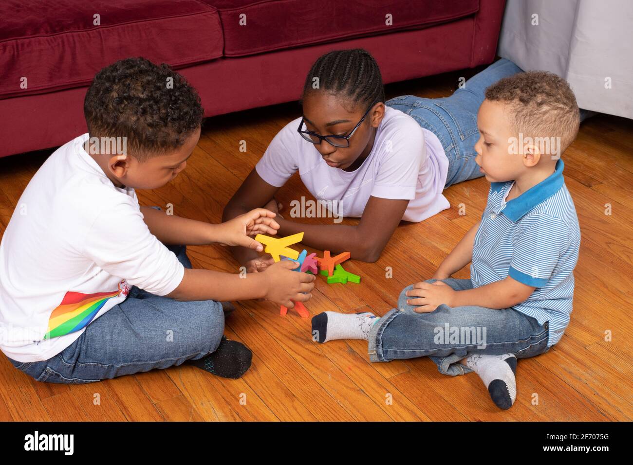 7 year old boy playing with wooden stacking pieces balancing game with 12 year old half-sister, 3 year old brother looking on learning by observing Stock Photo