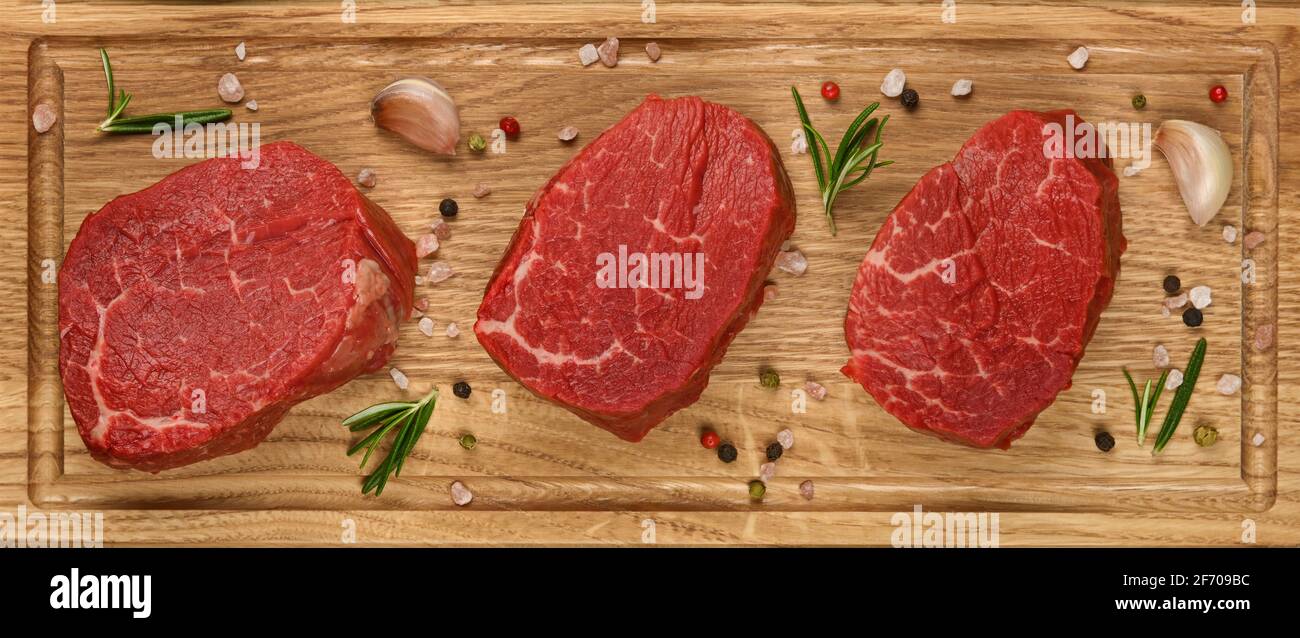 Close up three aged prime marbled raw tenderloin or fillet mignon beef steaks on brown oak wood cutting board, with spices, elevated top view, directl Stock Photo
