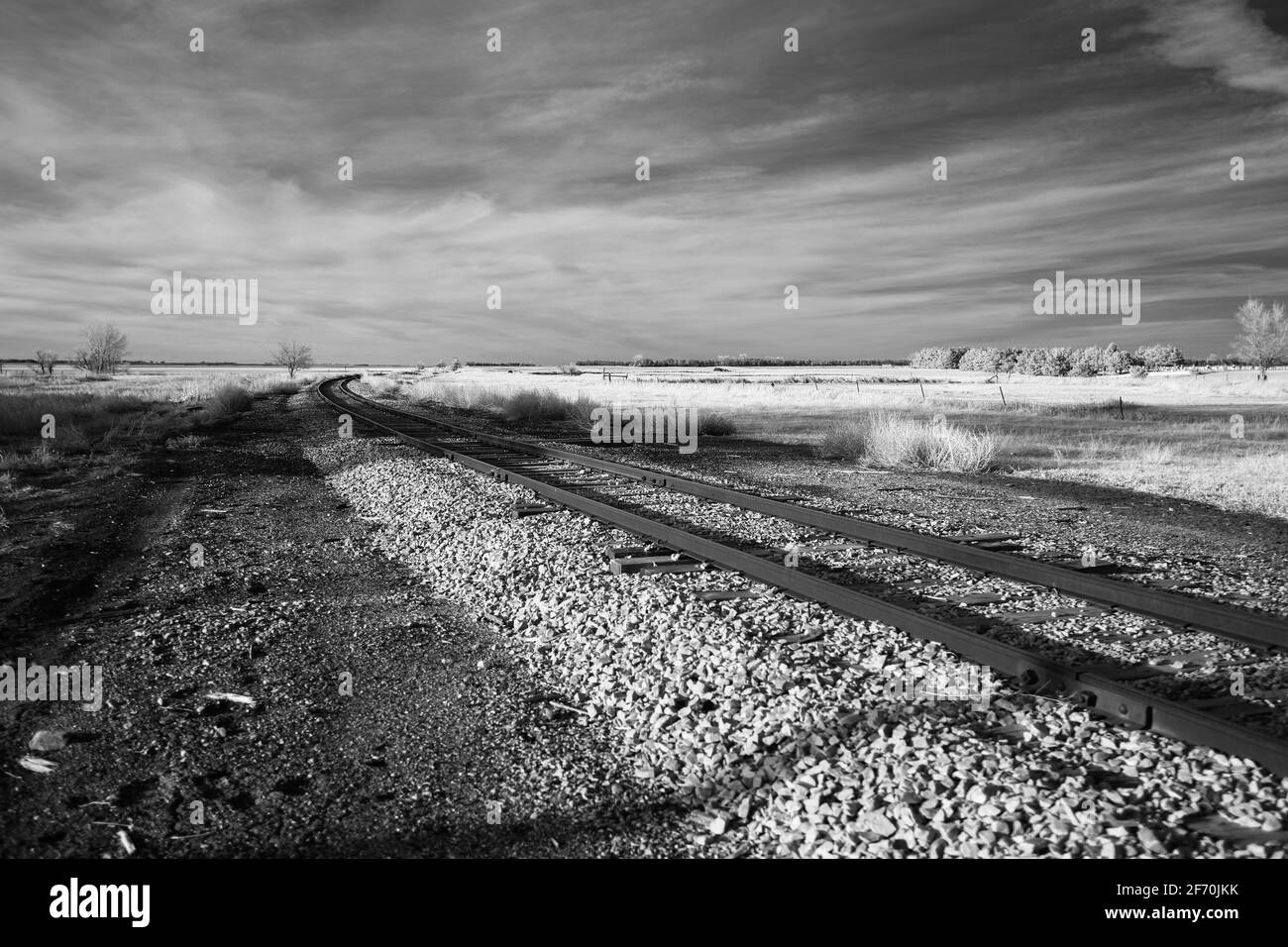 A curved set of train tracks vanishes behind a some brush in this black and white infrared image taken on the plains of South Dakota. Stock Photo