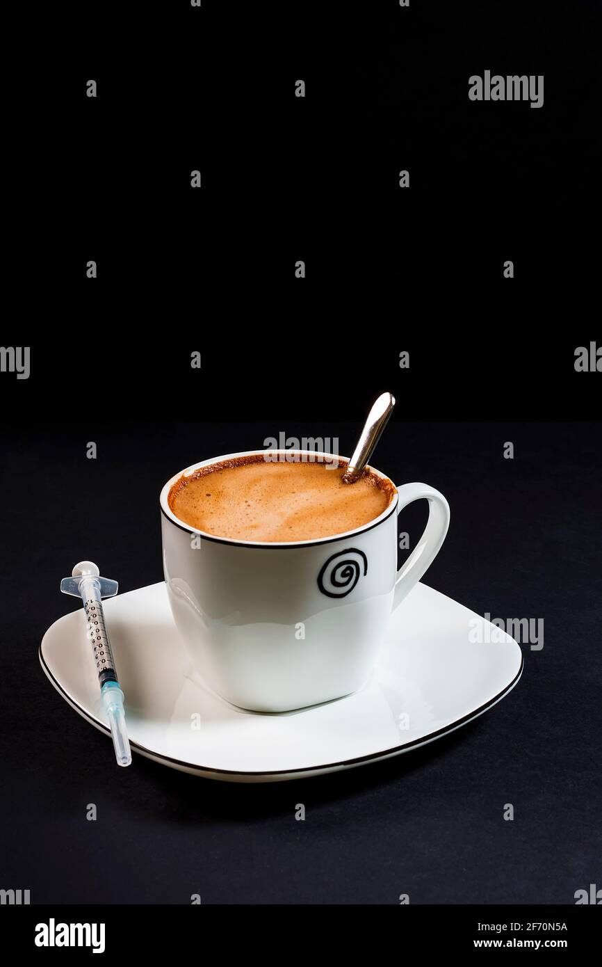 Coffee with milk in porcelain cup with syringe on saucer.This is a portrait format photo taken against a black background under artificial lighting in Stock Photo