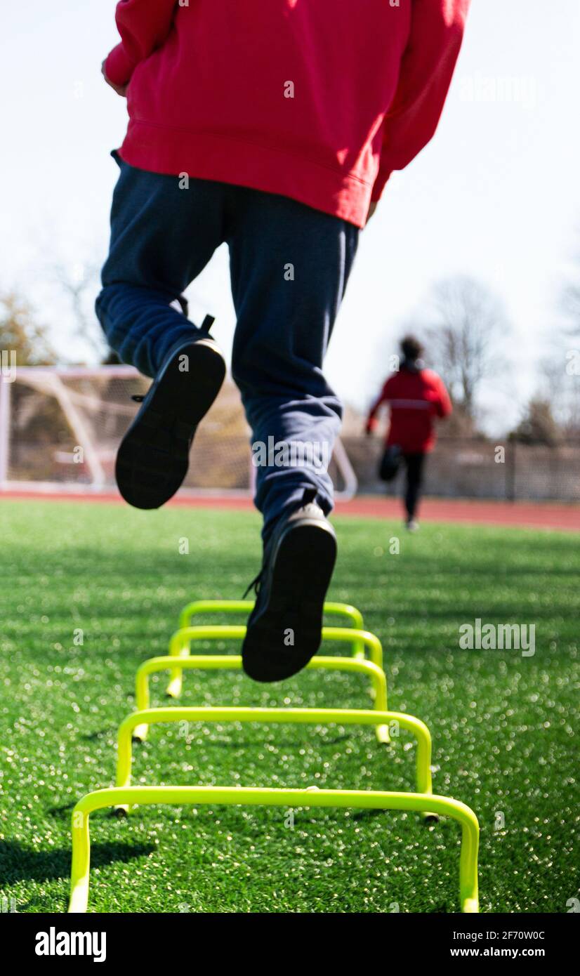 Rear view of a male athlete jumping on one leg over yellow mini hrdles on a gree turf field during track and field practice. Stock Photo