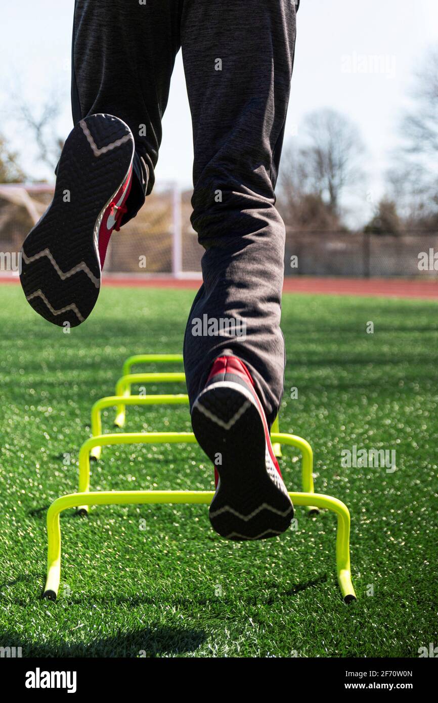 Rear view of a male athlete jumping on one leg over yellow mini hurdles on a green turf field during track and field practice from a low angle. Stock Photo