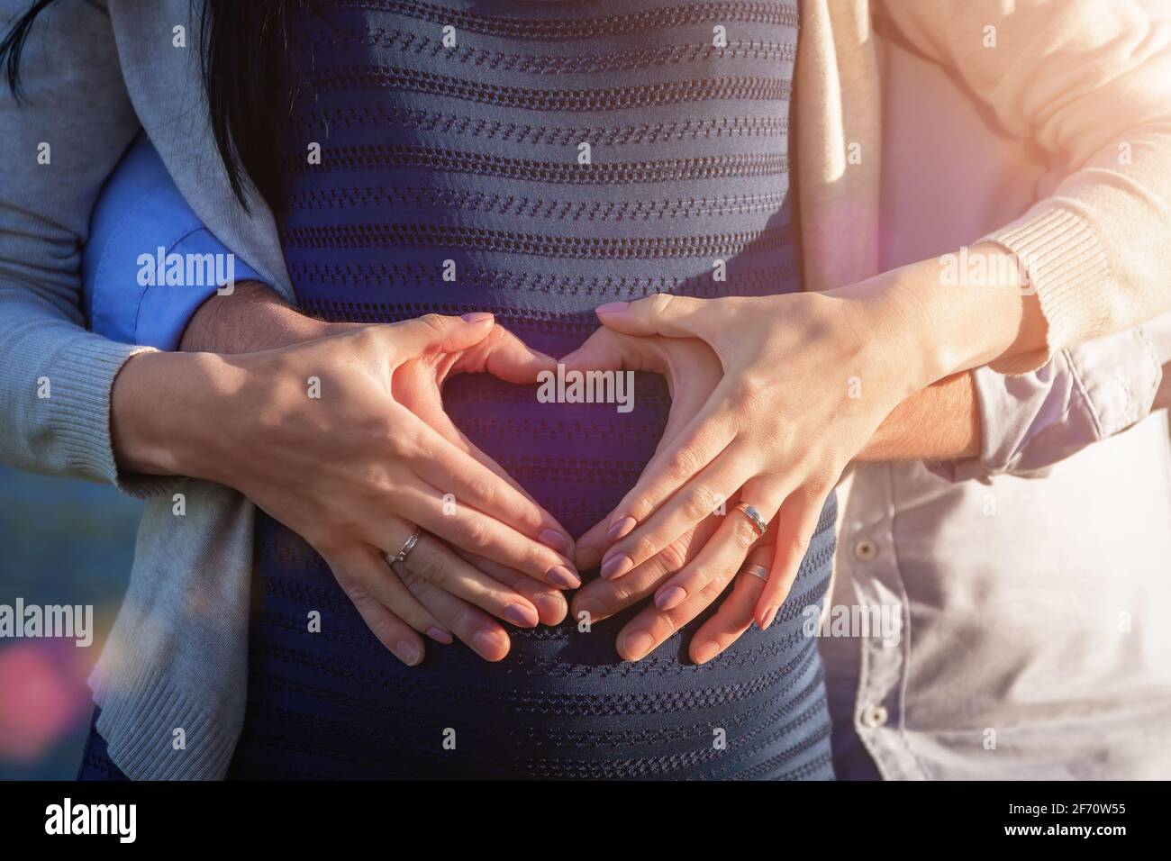 Hands of young couple at pregnant belly in heart shape symbol. Happy waiting for baby and motherhood. Stock Photo