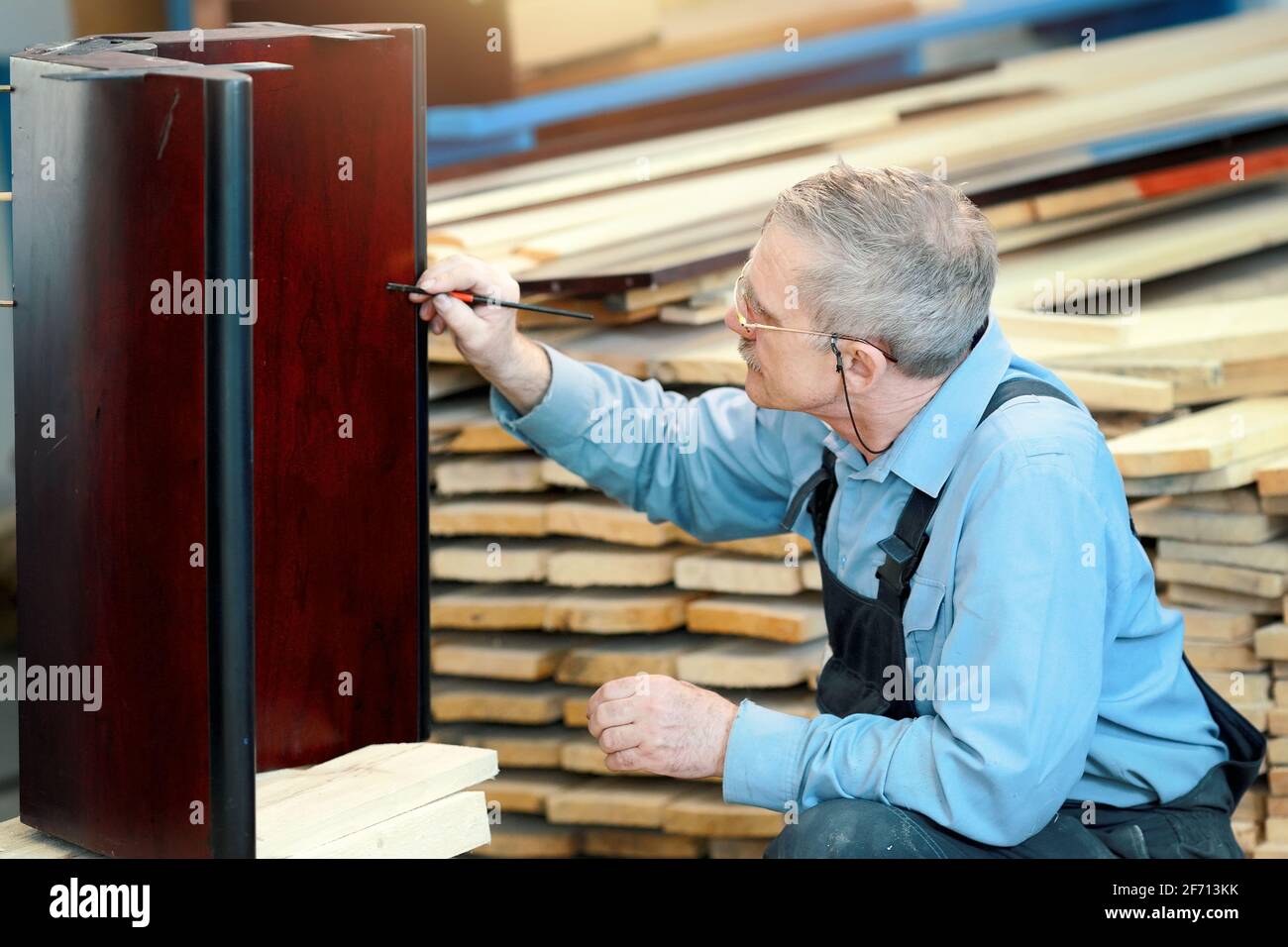 An elderly man with glasses and gray hair works with wood in a carpentry shop. A worker of Caucasian appearance is engaged in the restoration of furniture. Part-time job for a pensioner Stock Photo