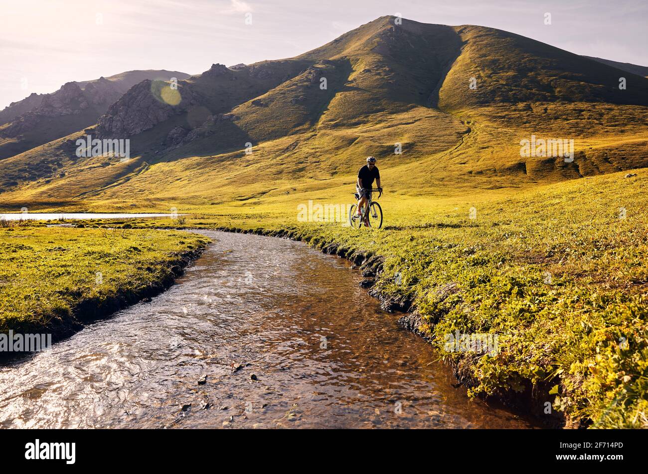 Man on mountain bike rides near the river at the green mountain valley at sunrise. Recreation, travel and healthy lifestyle concept. Stock Photo
