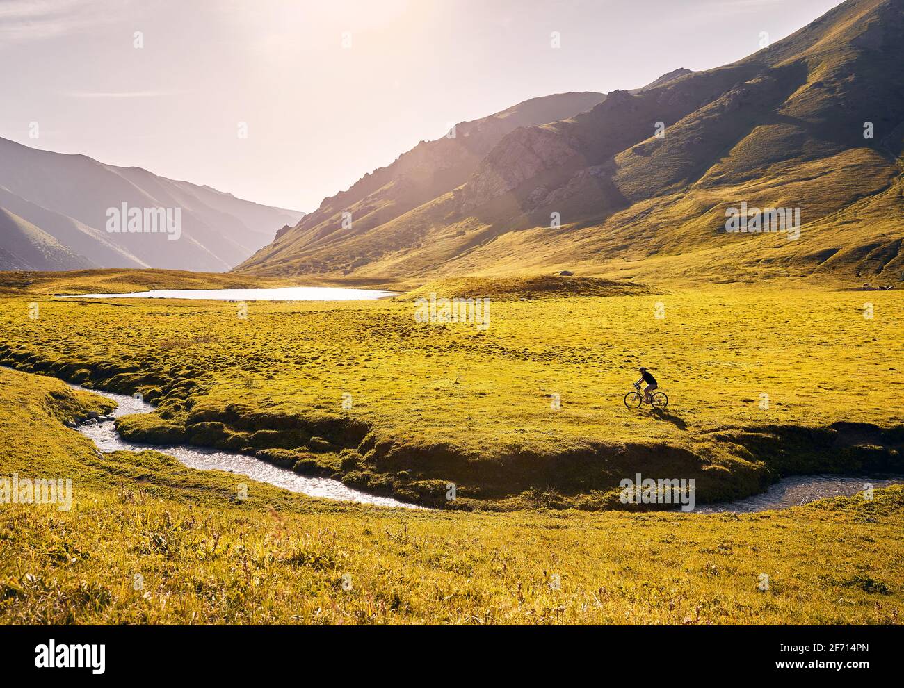 Man on mountain bike rides near the lake at the green mountain valley at sunrise. Recreation, travel and healthy lifestyle concept. Stock Photo
