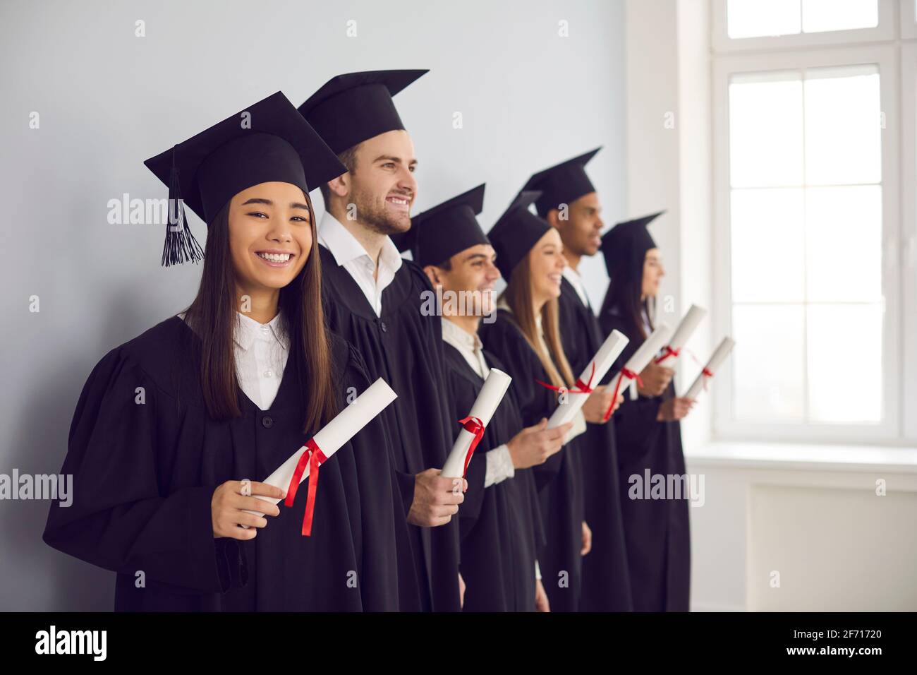 Side view of the university graduation class standing with diplomas in hand near the wall. Stock Photo