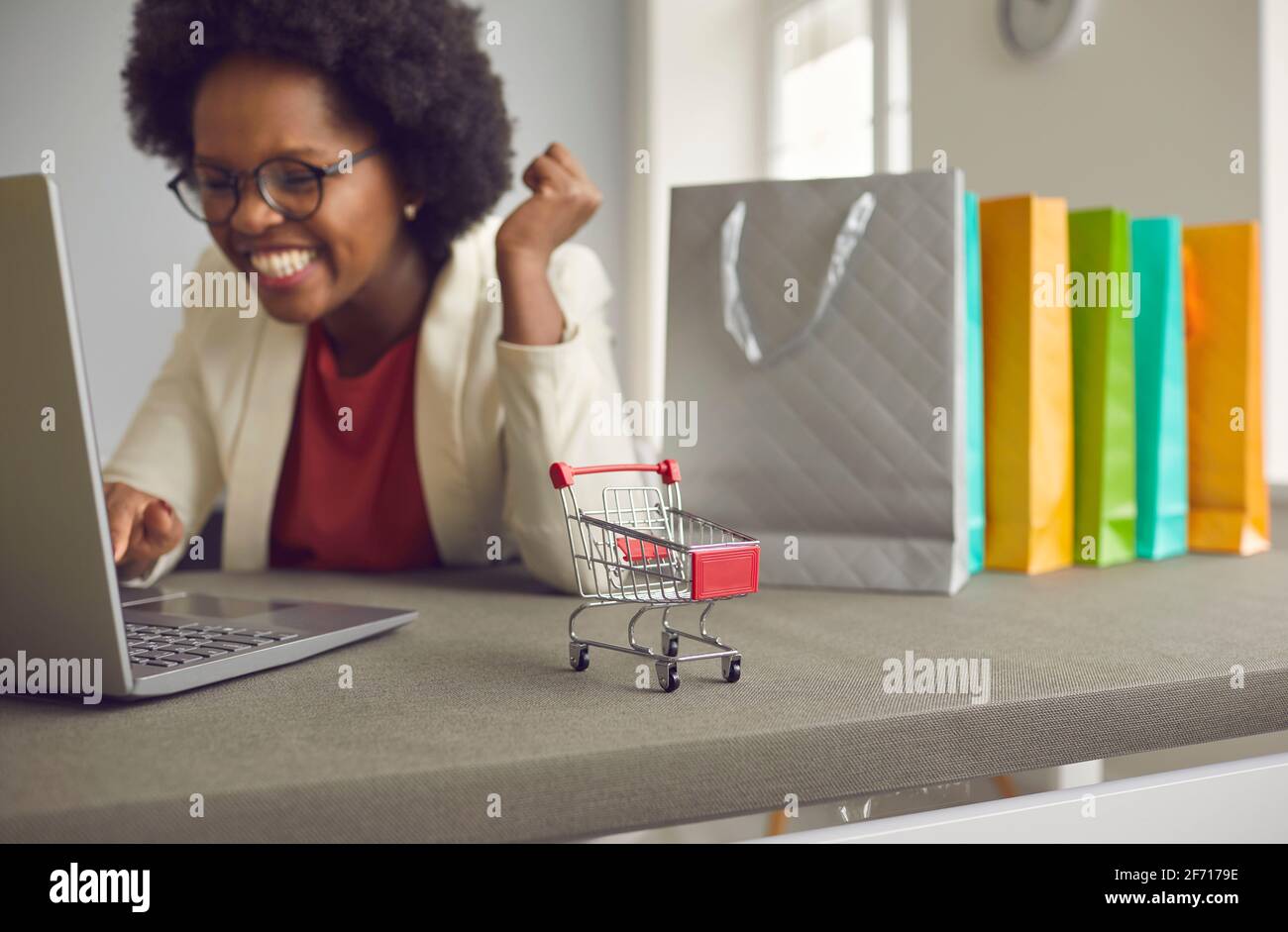 Close up of a small grocery cart on a table behind which a dark skinned woman is shopping online. Stock Photo