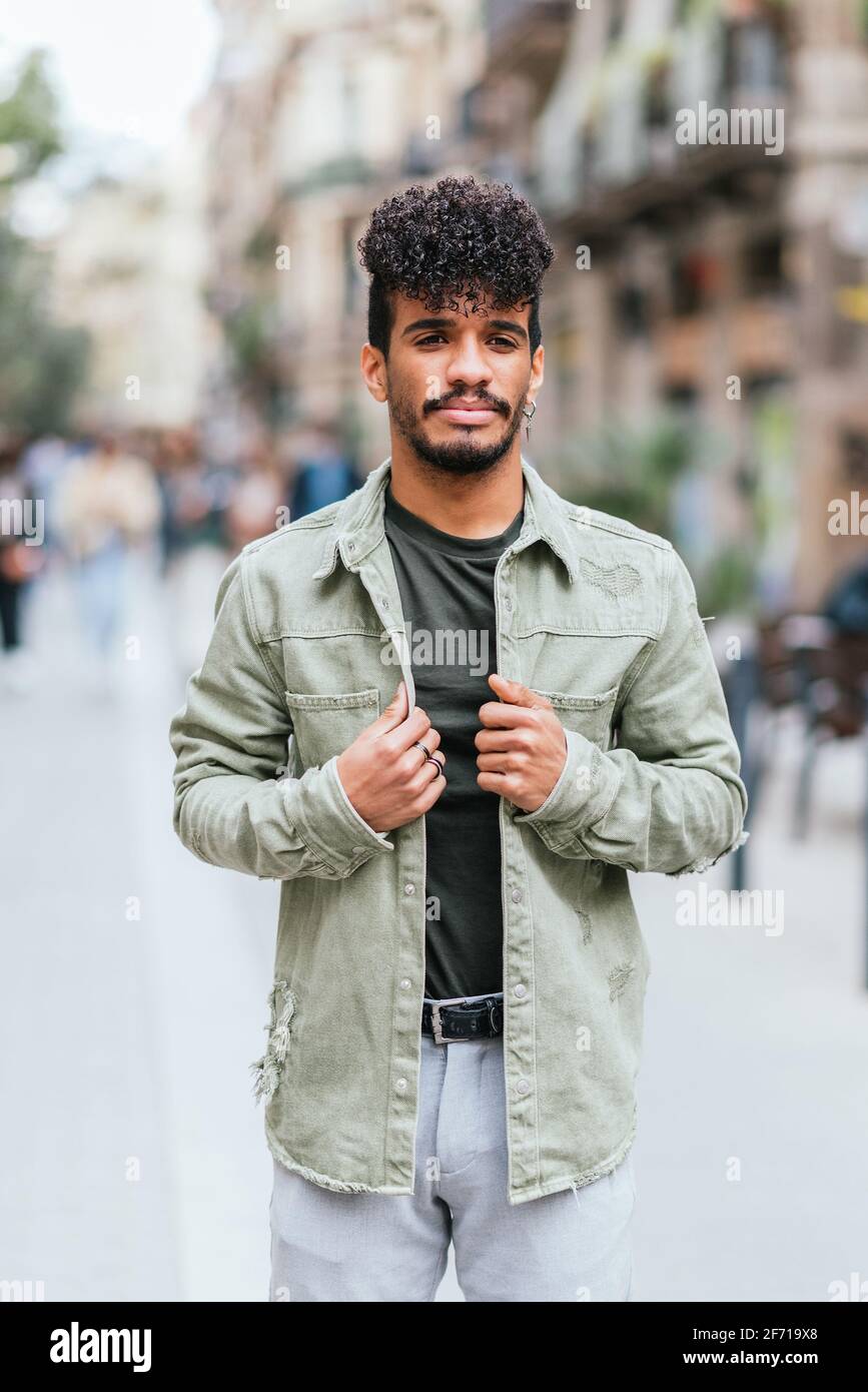 Vertical portrait of an hispanic young model posing in the streets of Barcelona. He is dressing casual green clothes and wears earrings and rings.  Stock Photo