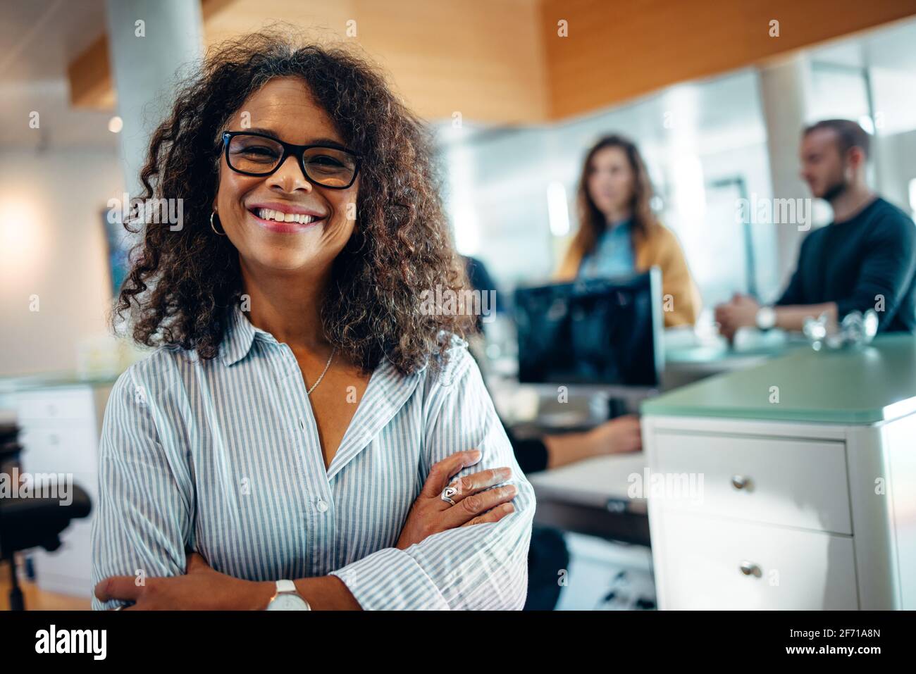 Portrait of a happy woman standing in a office with her arms crossed. Municipal office administrator looking at camera and smiling with people standin Stock Photo