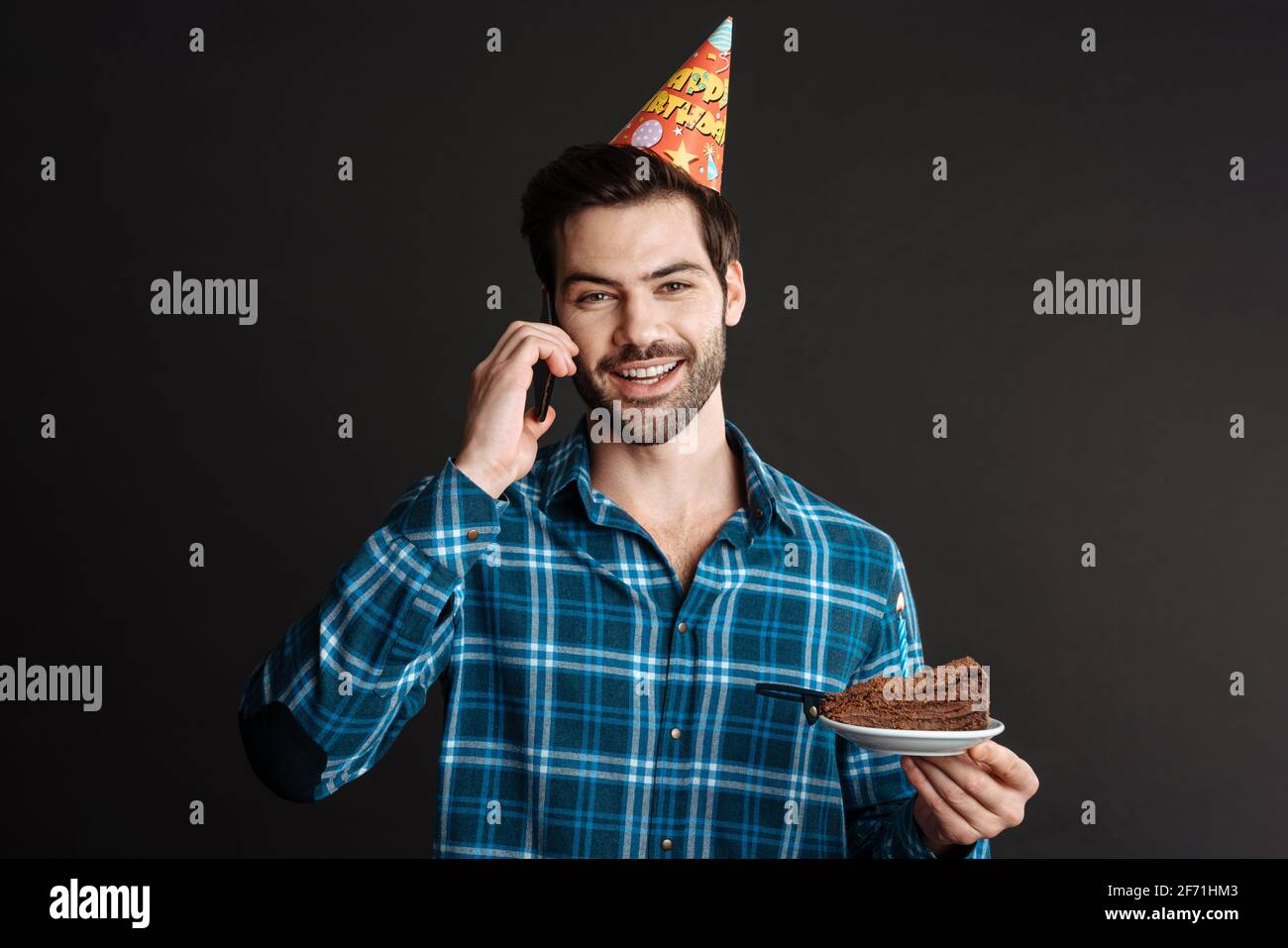 Smiling guy talking on cellphone while posing with birthday cake isolated over black background Stock Photo