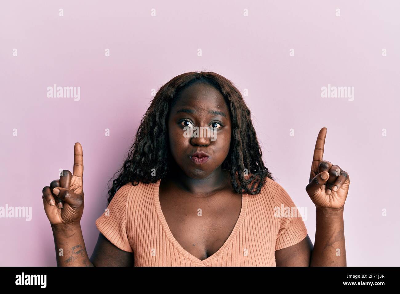African young woman pointing up with fingers puffing cheeks with funny face. mouth inflated with air, catching air. Stock Photo