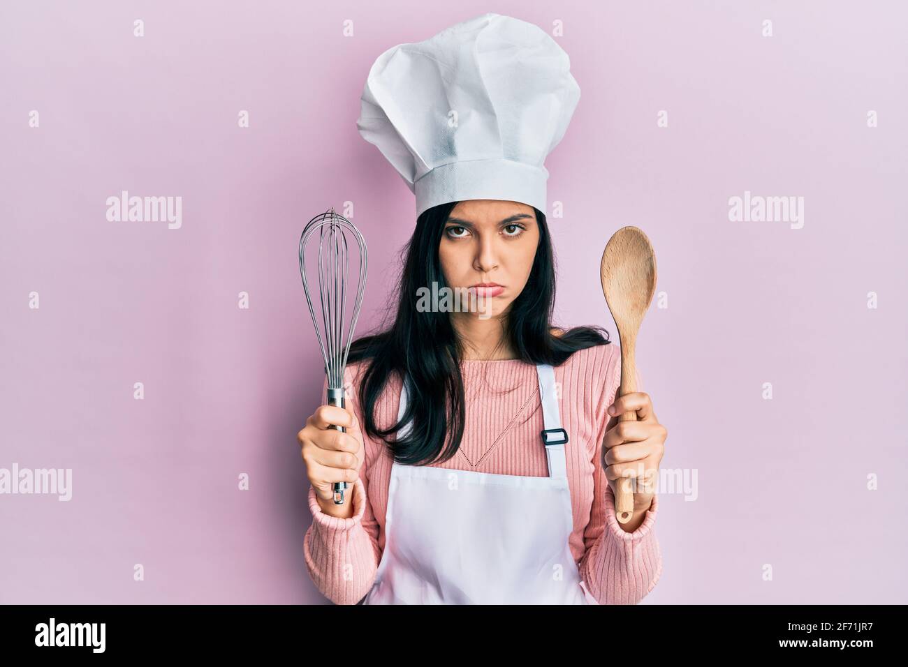 Young hispanic woman wearing baker uniform holding spoon and whisk ...