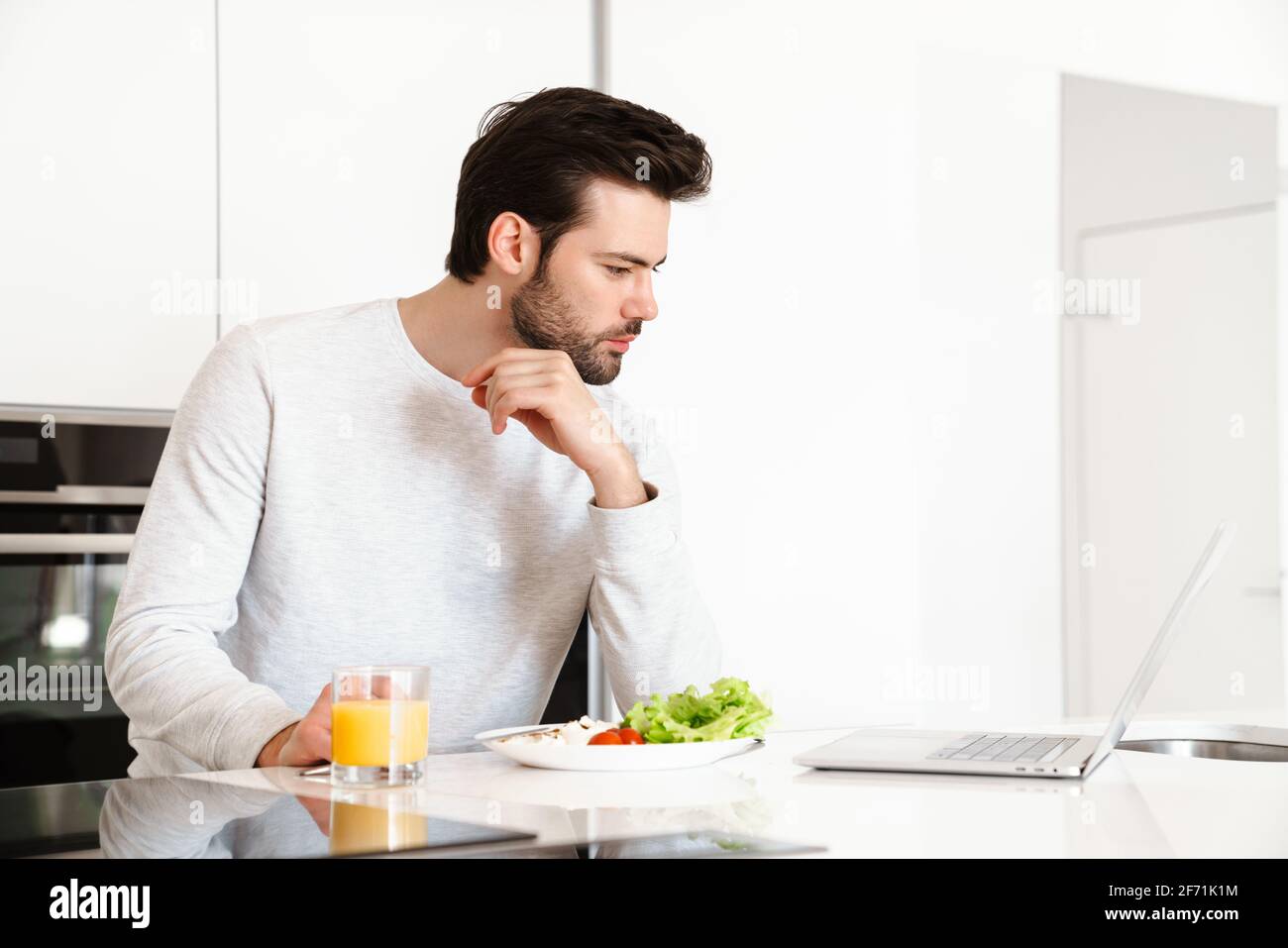 Focused handsome man using laptop while having breakfast at home kitchen Stock Photo