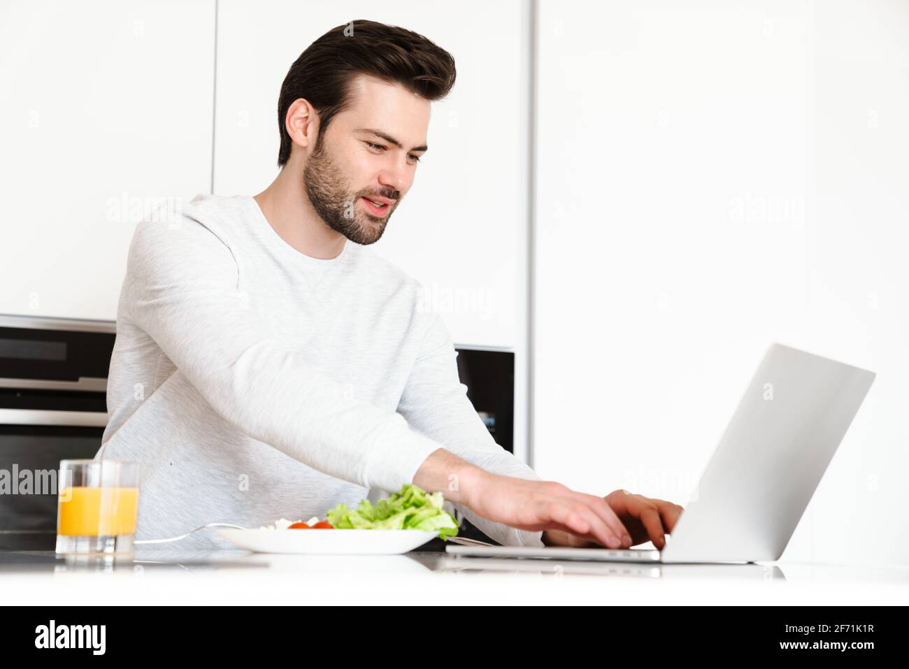 Pleased handsome man using laptop while having breakfast at home kitchen Stock Photo