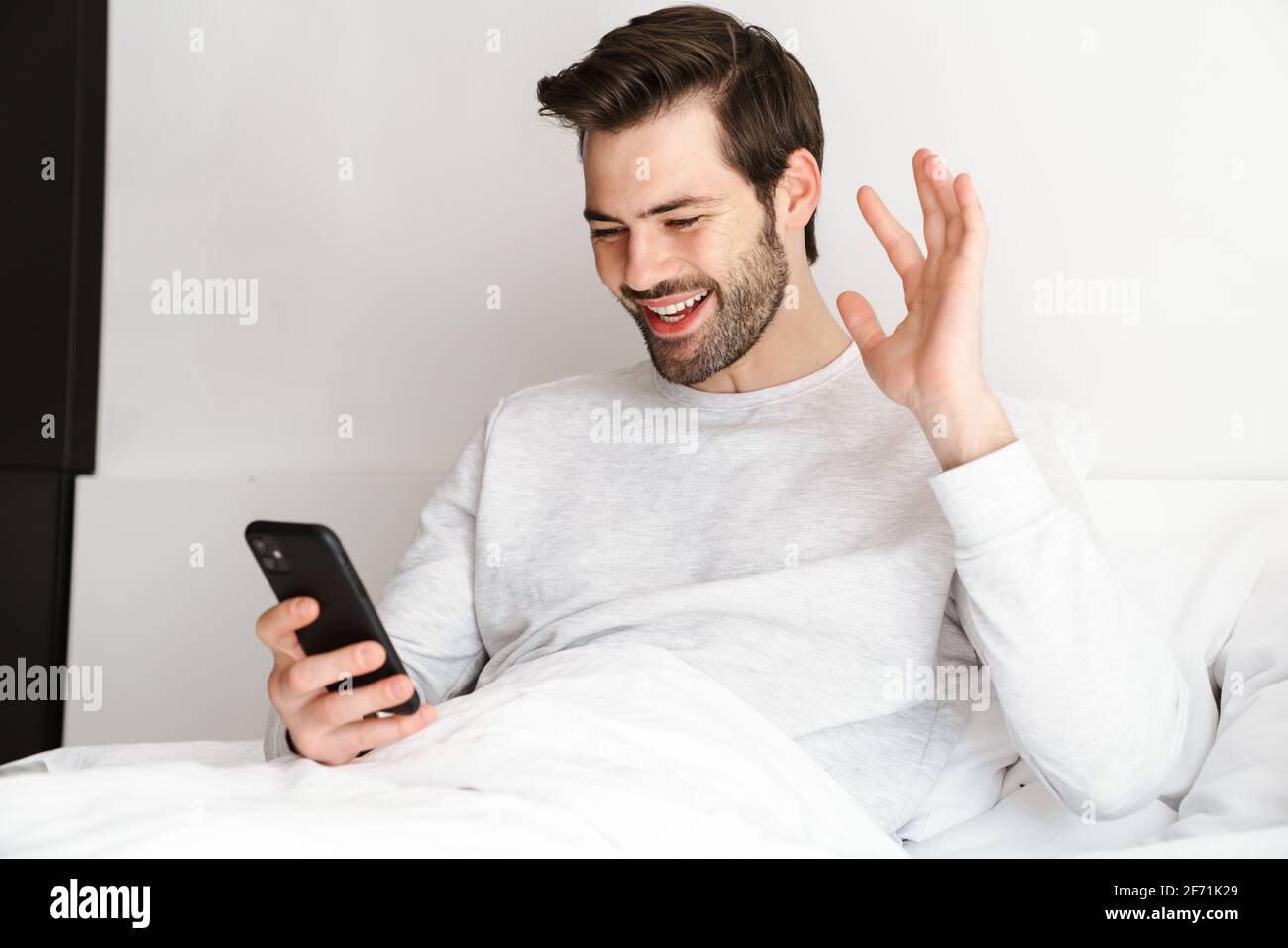 Smiling man waving hand and using mobile phone while resting in bed at home Stock Photo