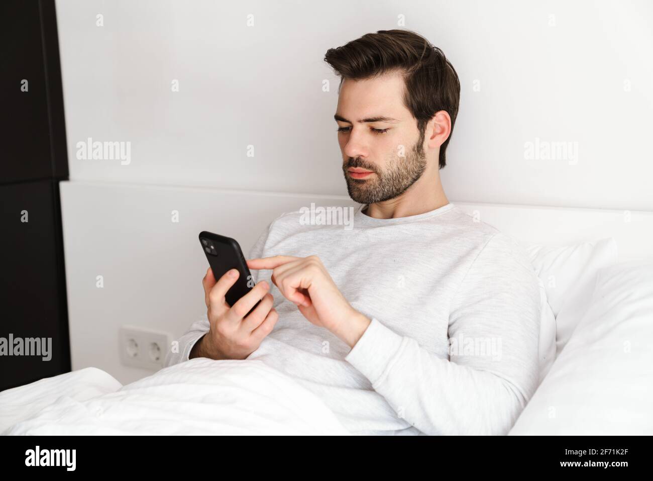 Focused handsome man using mobile phone while resting in bed at home Stock Photo