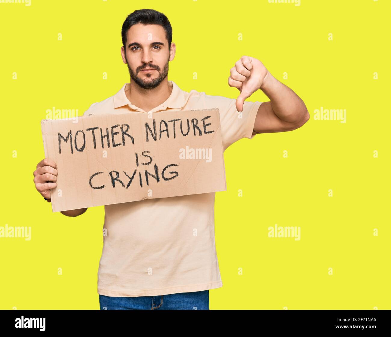 Young hispanic man holding mother nature is crying protest cardboard banner with angry face, negative sign showing dislike with thumbs down, rejection Stock Photo
