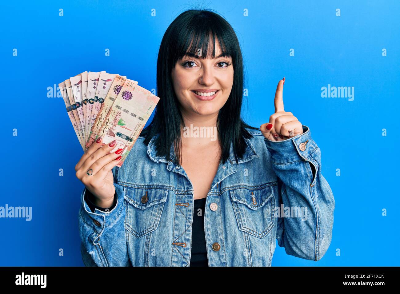 Young hispanic woman holding saudi arabia riyal banknotes smiling with an idea or question pointing finger with happy face, number one Stock Photo