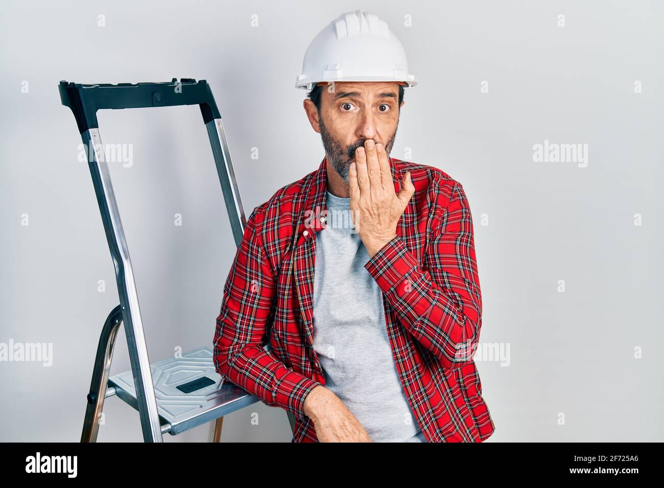 Middle age hispanic worker man using ladder covering mouth with hand ...