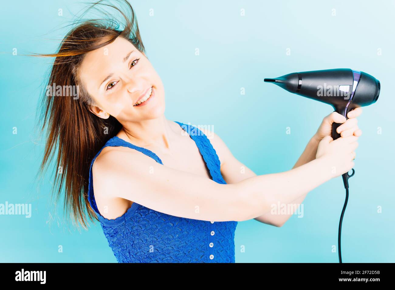 Beautiful young woman drying her hair with blow dryer smiling with blue ...