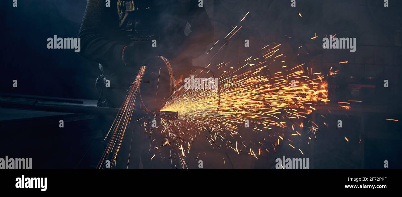 Close up of young man in special dark suit grinding metal on steel pipe with flash sparks. Concept of process working with angle grinder and metal in factory or garage. Stock Photo