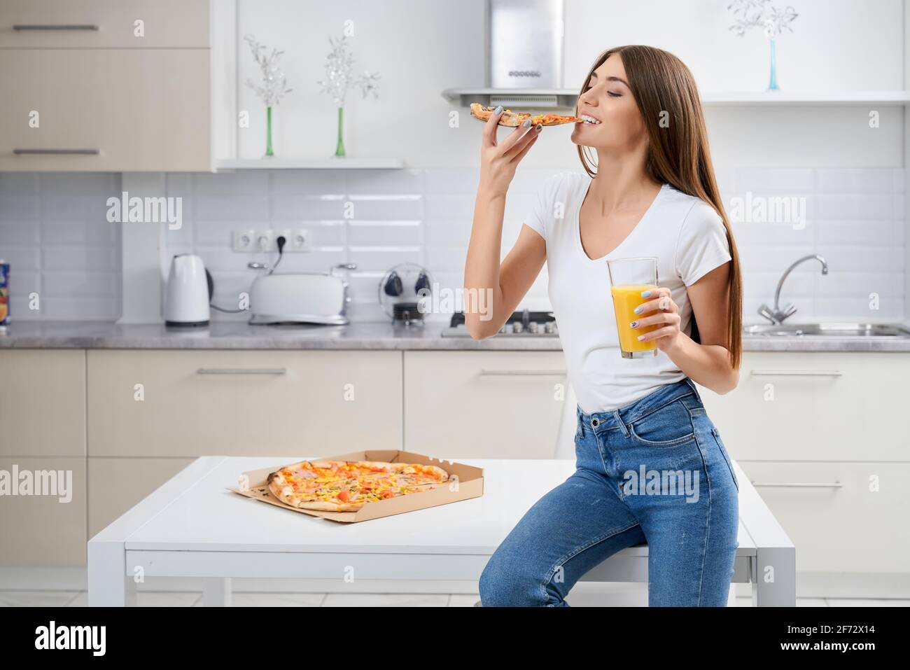 Beautiful brunette sitting on table and enjoying tasty pizza and drinking juice in kitchen. Concept of tasty food. Stock Photo