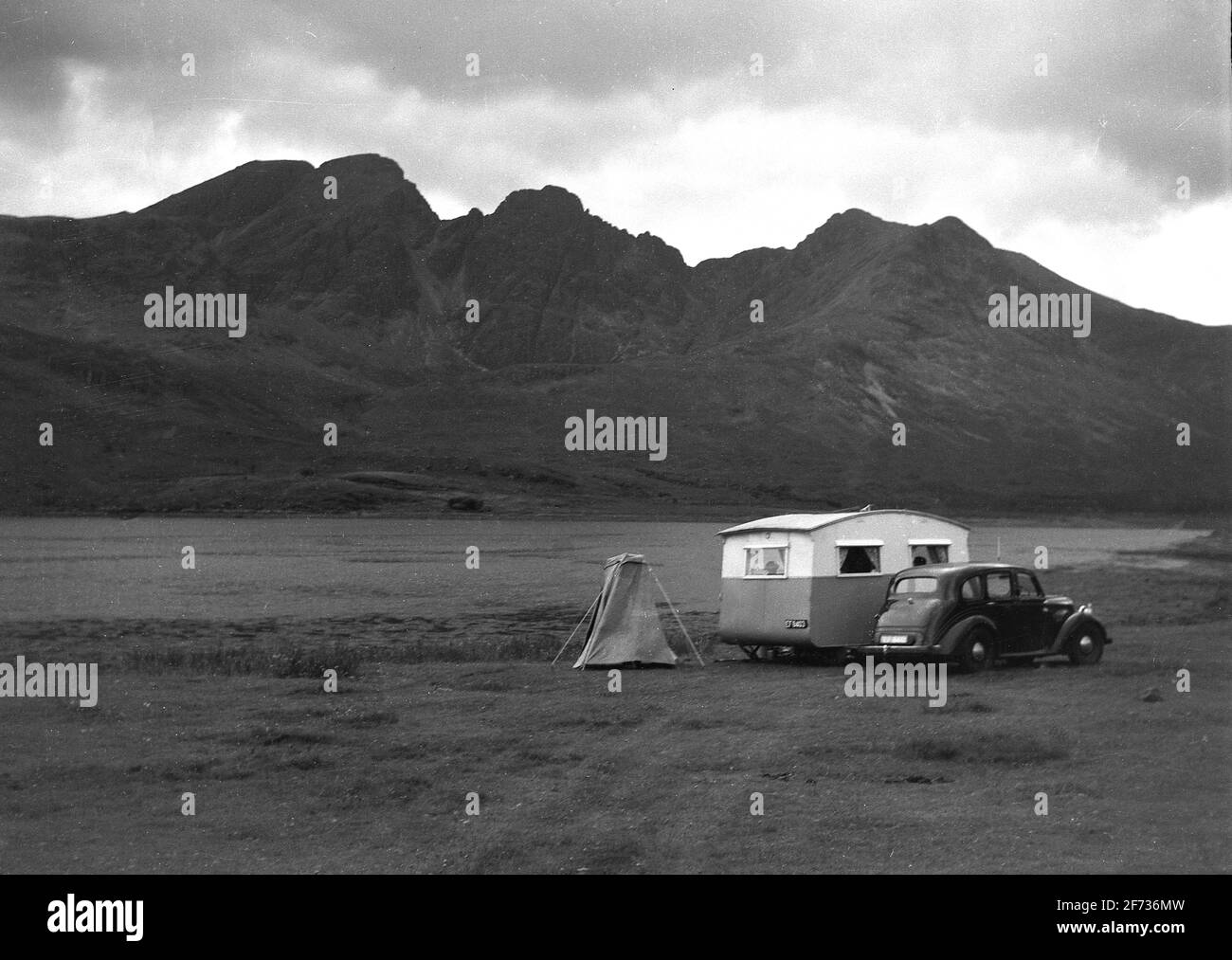 1956, historical, a motoring holiday in the Scottish highlands. A motor car and caravan of the era parked in a wide open space  or lowland, with a mountain range in the distance. A small tent sits beside the caravan. The remote area is famous for its beautiful scenery and mountainous landscape, with the region being the location of the UK's highest peak, 'Ben Nevis.'. Stock Photo