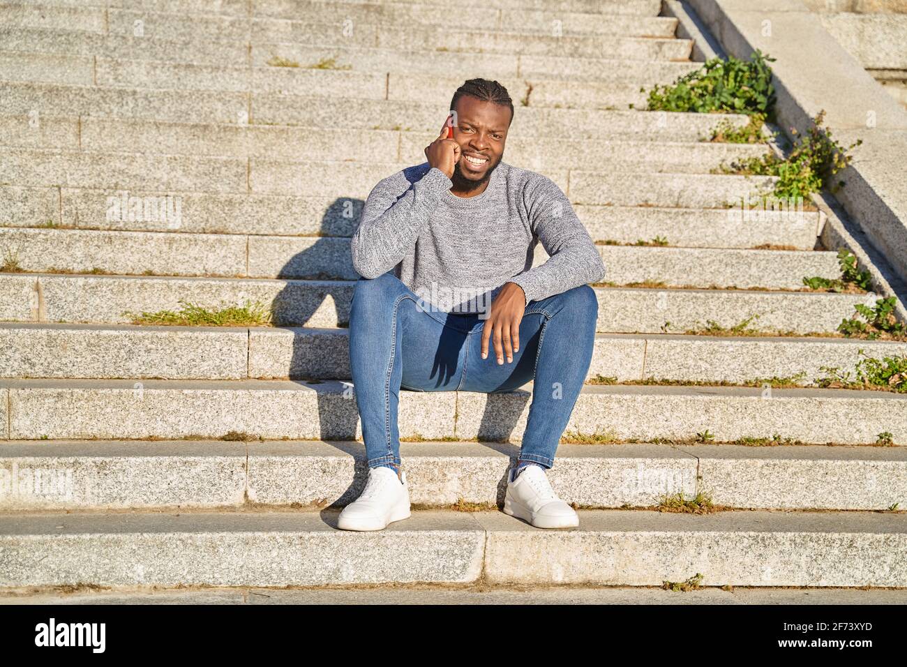 Happy black man talking on smart phone sitting on stairs. Young man in jeans and grey sweater. Concept of communication. High quality photo Stock Photo