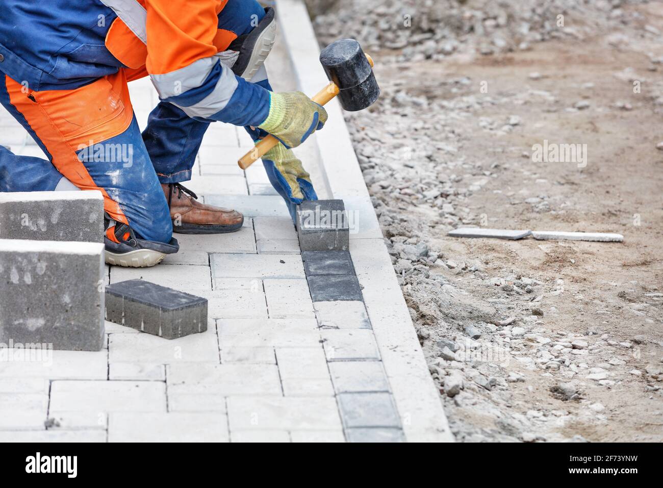 A worker in blue and orange overalls, half-squatting, taps the paving slabs with a rubber mallet. Close-up, selective focus, copy space. Stock Photo