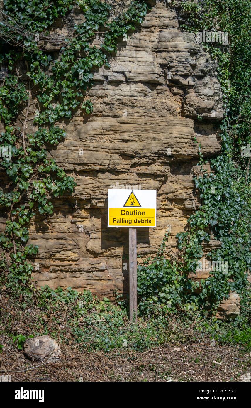 Beware of falling rock warning sign with triangle picture and alert message in yellow and black.  Stood in front of cliff face with rock on the ground Stock Photo