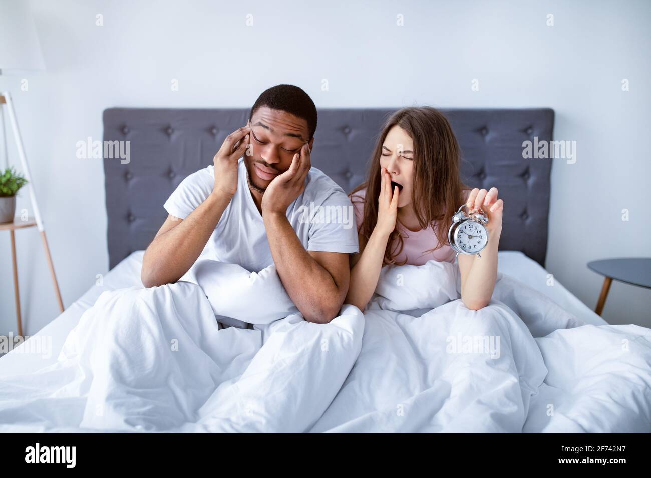 Multiracial young couple feeling too tired to wake up, feeling sleepy, holding alarm clock, being late to work Stock Photo