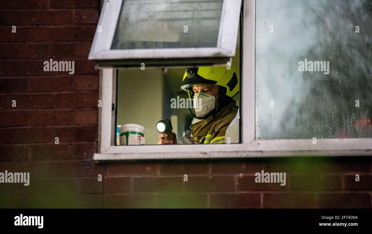 British firefighter wearing white face mask and carrying torch seen through the window of a house where a fire took place. He is investigating the cause of the fire in a smokey room. Stock Photo