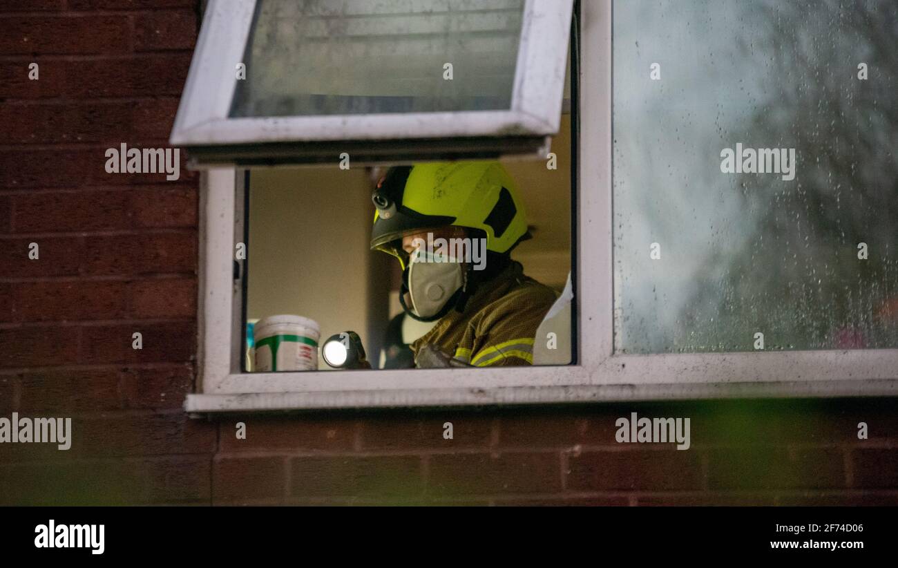 British firefighter wearing white face mask and carrying torch seen through the window of a house where a fire took place. He is investigating the cause of the fire in a smokey room. Stock Photo