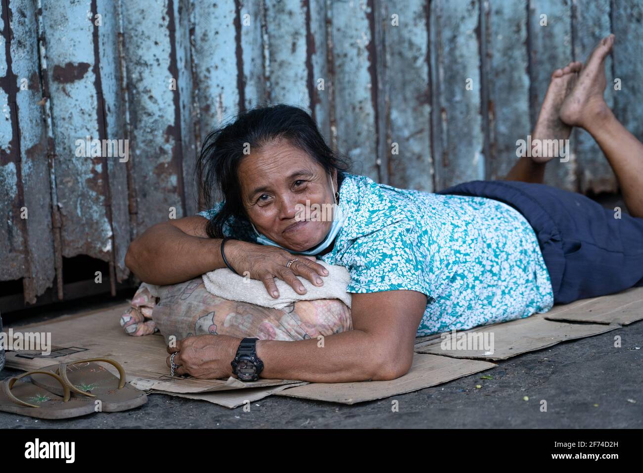A homeless Filipino woman smiles towards the camera whilst lying on the side street on cardboard where she sleeps Stock Photo