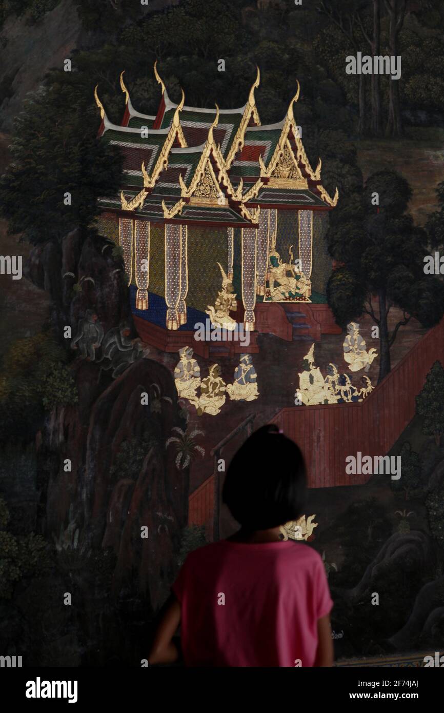 A visitor paying attention to a mural painting, part of a gallery of mural paintings depicting Ramakien (Thai version of Ramayana epic) which is located at the Wat Phra Kaew (Temple of Emerald Buddha), inside Grand Palace complex in Bangkok, Thailand. Thailand is the first Southeast Asian country to reduce quarantine restrictions--from 14 days to 10 days--to speed up the recovery of its tourism sector, according to South China Morning Post on April 2, 2021. The decision was made to speed up the recovery process, that was previously forecasted by World Bank to be 'a slow return.' Stock Photo