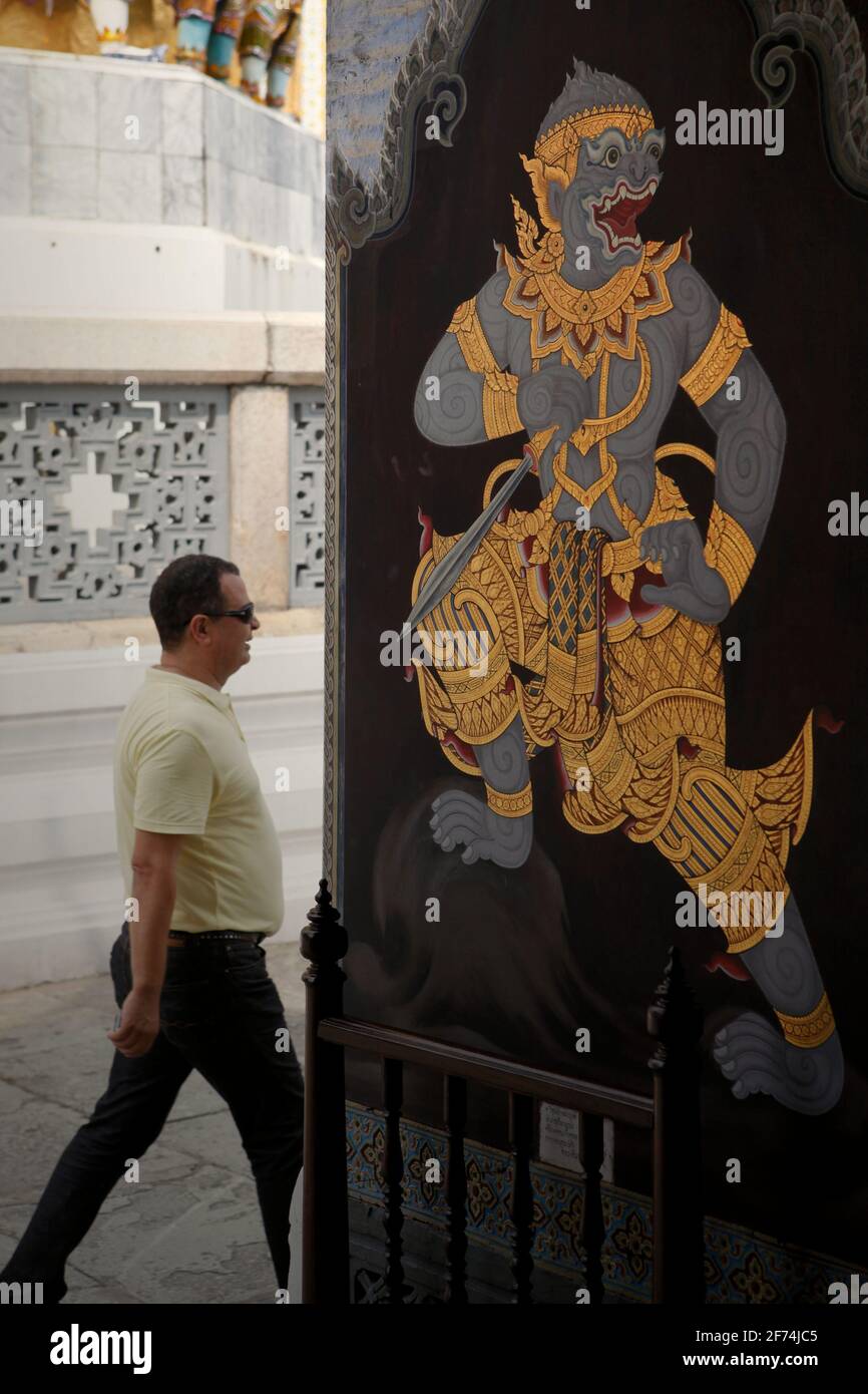 A visitor walking pass an entrance where a mural painting is placed on its wall, part of a gallery of mural paintings depicting Ramakien (Thai version of Ramayana epic) which is located at the Wat Phra Kaew (Temple of Emerald Buddha), inside Grand Palace complex in Bangkok, Thailand. Thailand is the first Southeast Asian country to reduce quarantine restrictions--from 14 days to 10 days--to speed up the recovery of its tourism sector, according to South China Morning Post on April 2, 2021. The decision was made to speed up the recovery process, that was forecasted to be 'a slow return.' Stock Photo