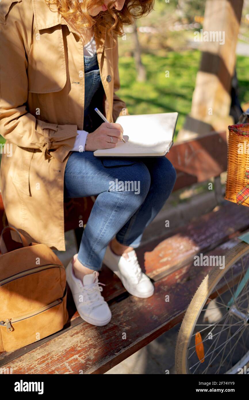 Crop of anonymous young thoughtful female taking notes in planner on wooden bench near bicycle in park in daytime Stock Photo