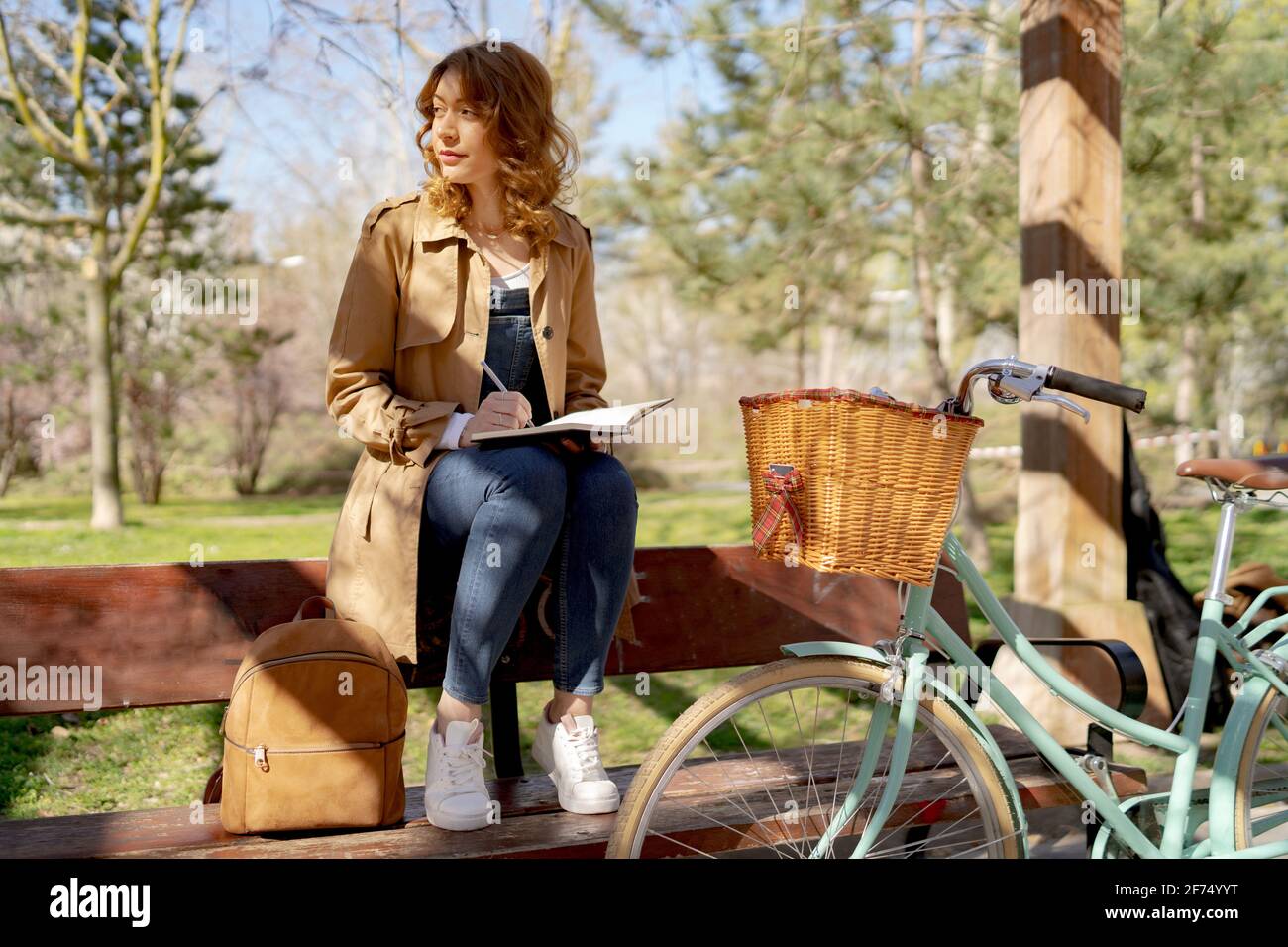 Young thoughtful female taking notes in planner on wooden bench near bicycle in park in daytime Stock Photo