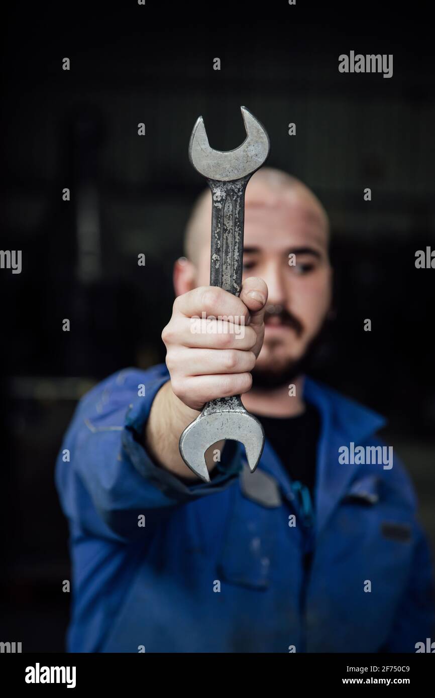 Male mechanic in blue overall covering face with metal spanner in hands looking away on black background Stock Photo