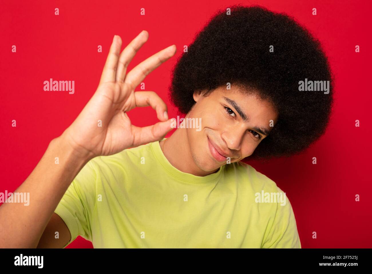 Positive African American male with Afro hairstyle smiling and looking at camera and showing okay sign while standing on red background Stock Photo
