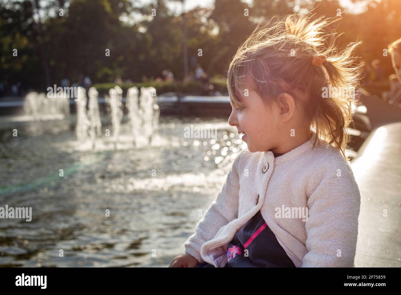 adorable toddler girl looks at the pond with fountains in the park on a sunny day. weekend family walk. spending time with children. artistic focus Stock Photo