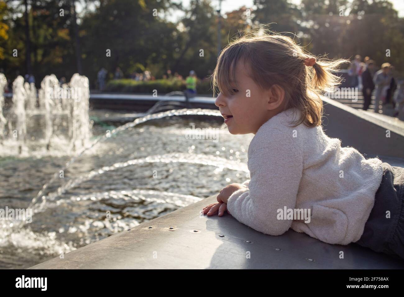 adorable toddler girl looks at the pond with fountains in the park on a sunny day. weekend family walk. spending time with children. artistic focus Stock Photo
