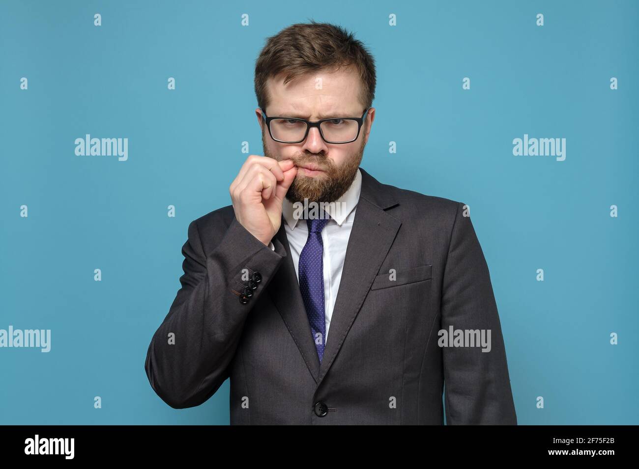 Suspicious Caucasian man in business suit, sternly and suspiciously looks, touching mustache with hands. Blue background. Stock Photo
