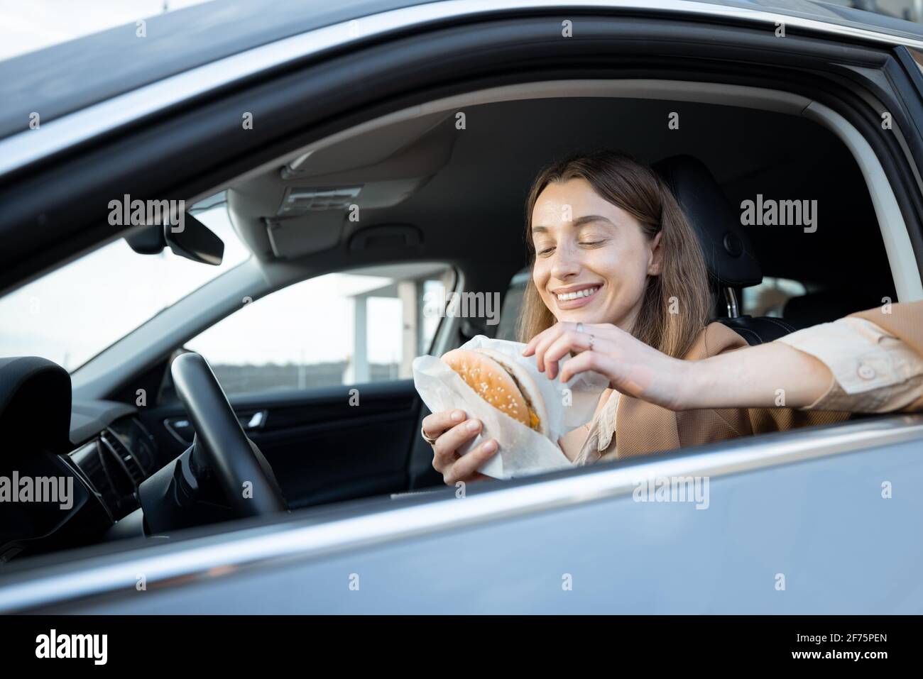 Happy woman eating a burger in the car. Have unhealthy fast food snack. Food to go. Hungry and busy concept. Stock Photo