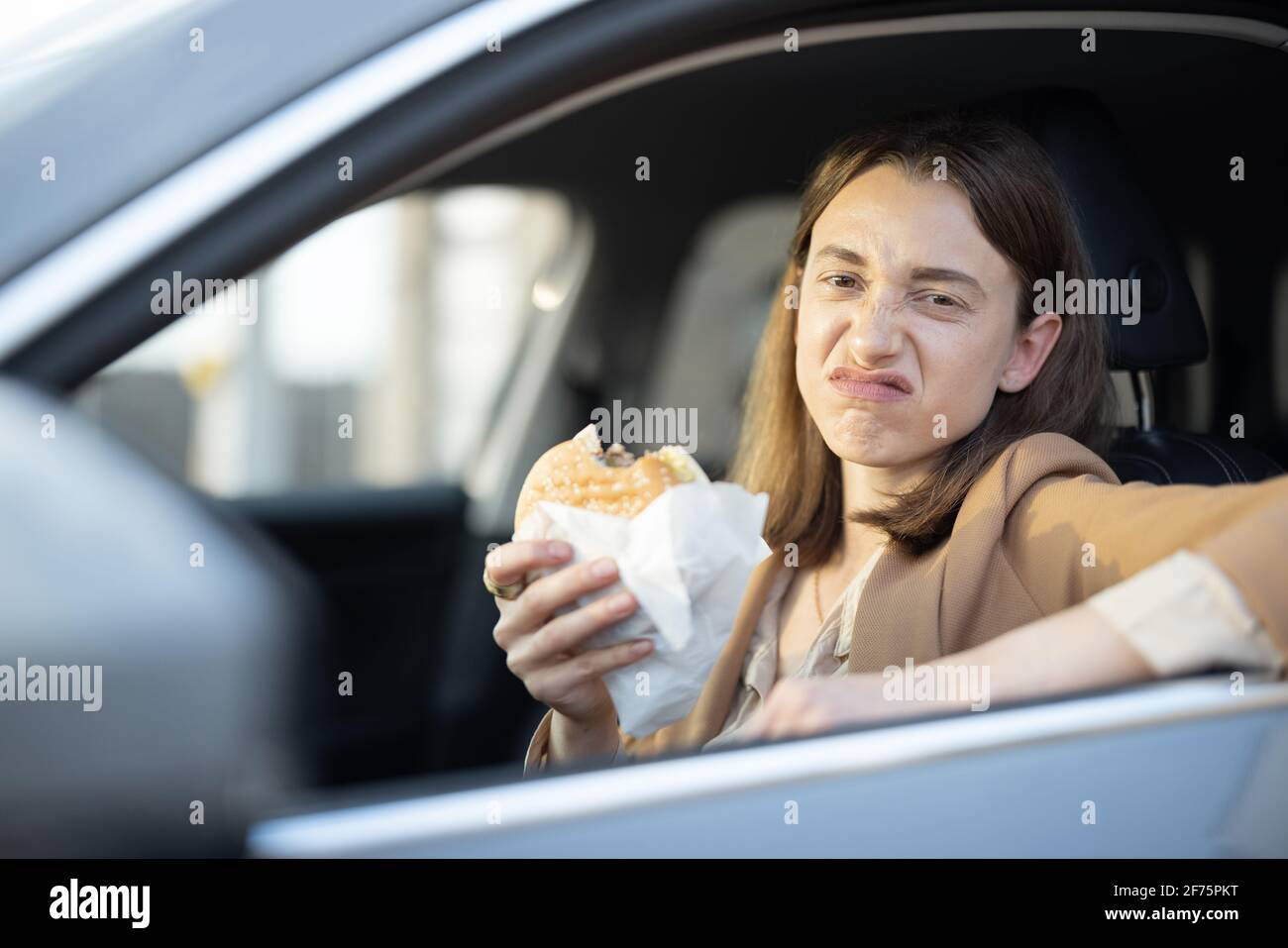 Grimaced woman eating a burger in the car. Have unhealthy tasteless fast food snack. Food to go. Hungry and busy concept. Stock Photo