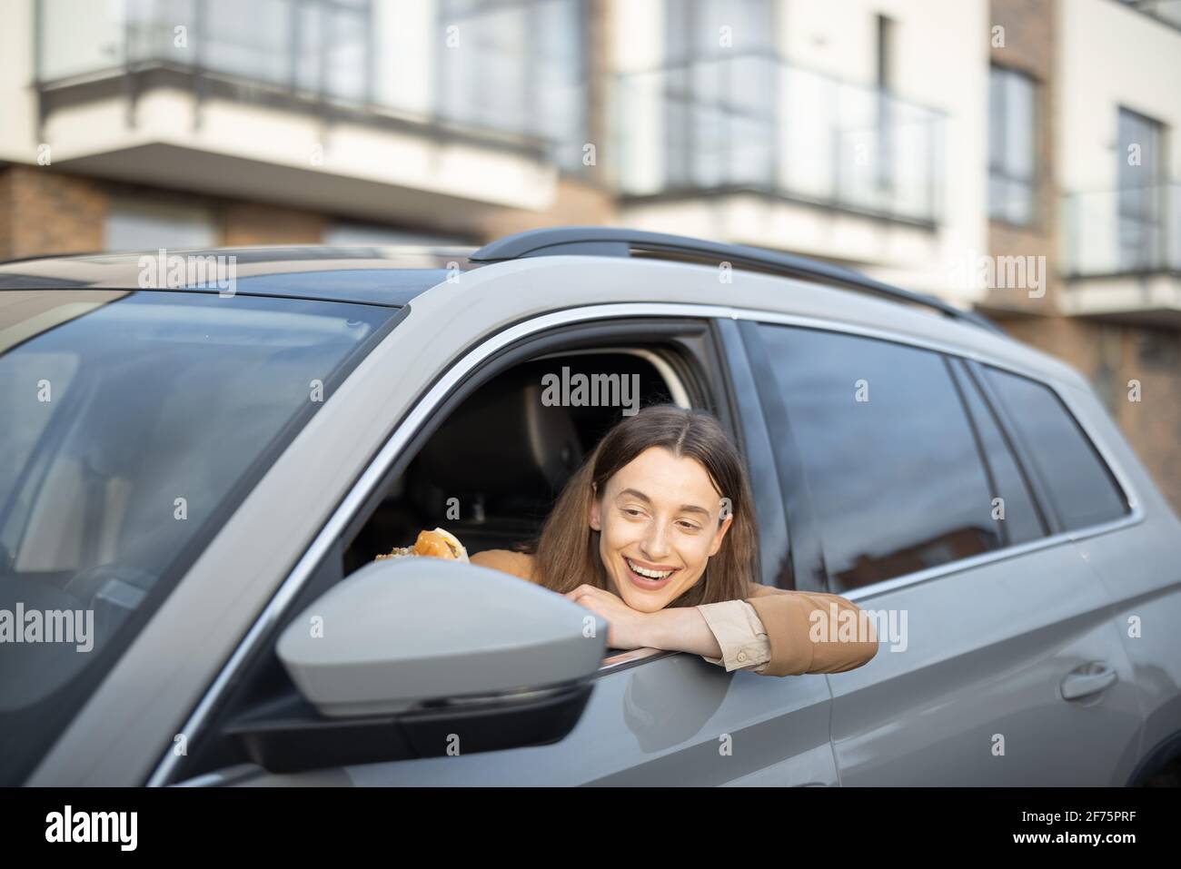 Happy woman eating a burger in the car. Have unhealthy fast food snack. Food to go. Hungry and busy concept. Stock Photo