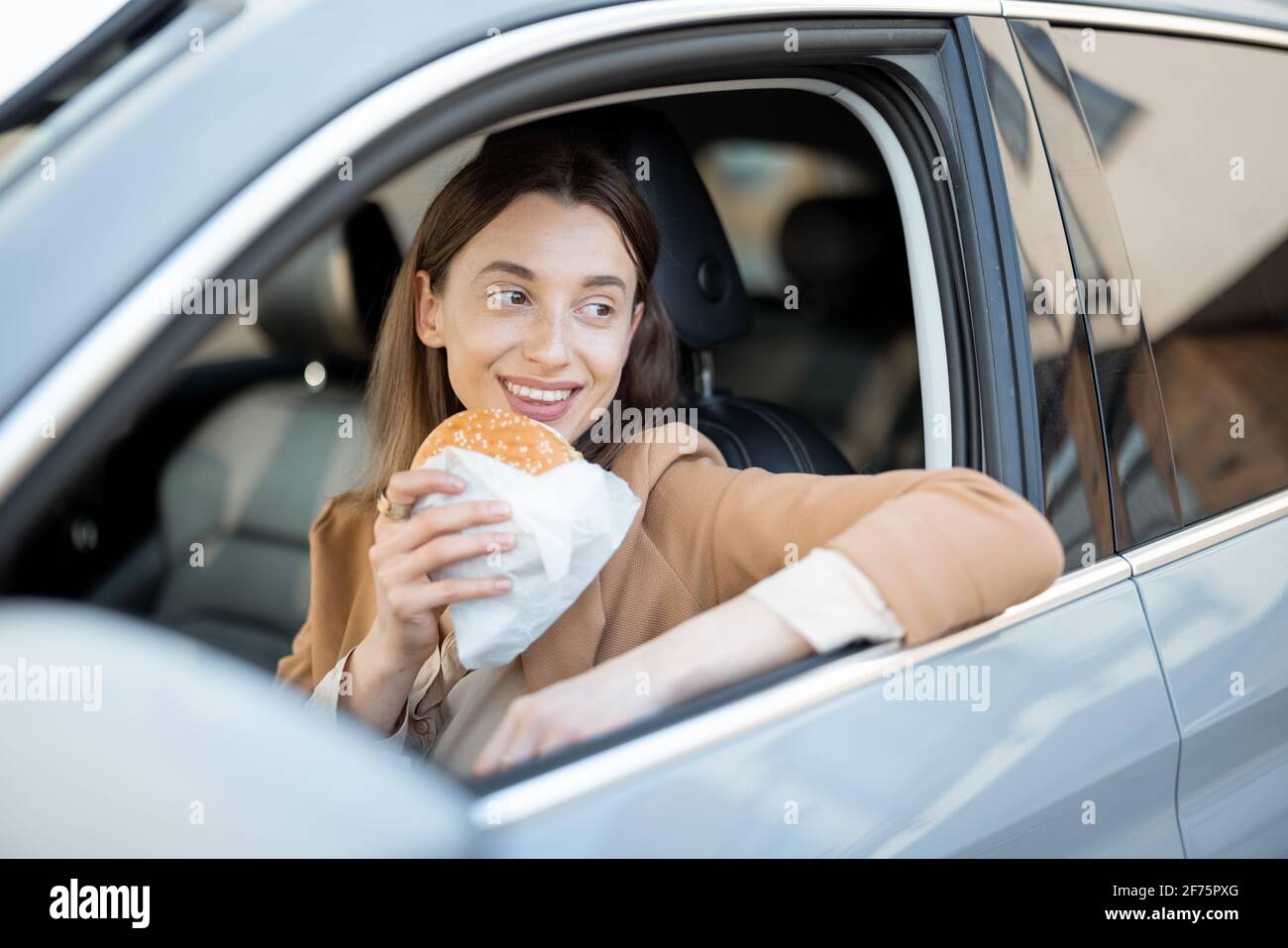 Happy woman eating a burger in the car. Have unhealthy fast food snack. Food to go. Hungry and busy concept. Stock Photo